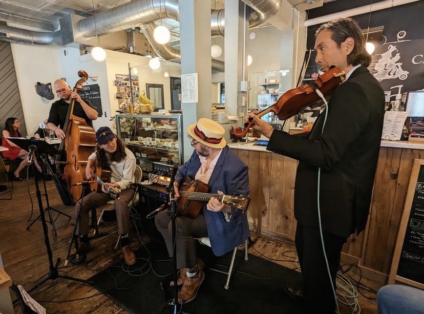 Image shows four musicians making eye contact with one another as they perform in a cafe with their double-bass, two guitars, and a violin